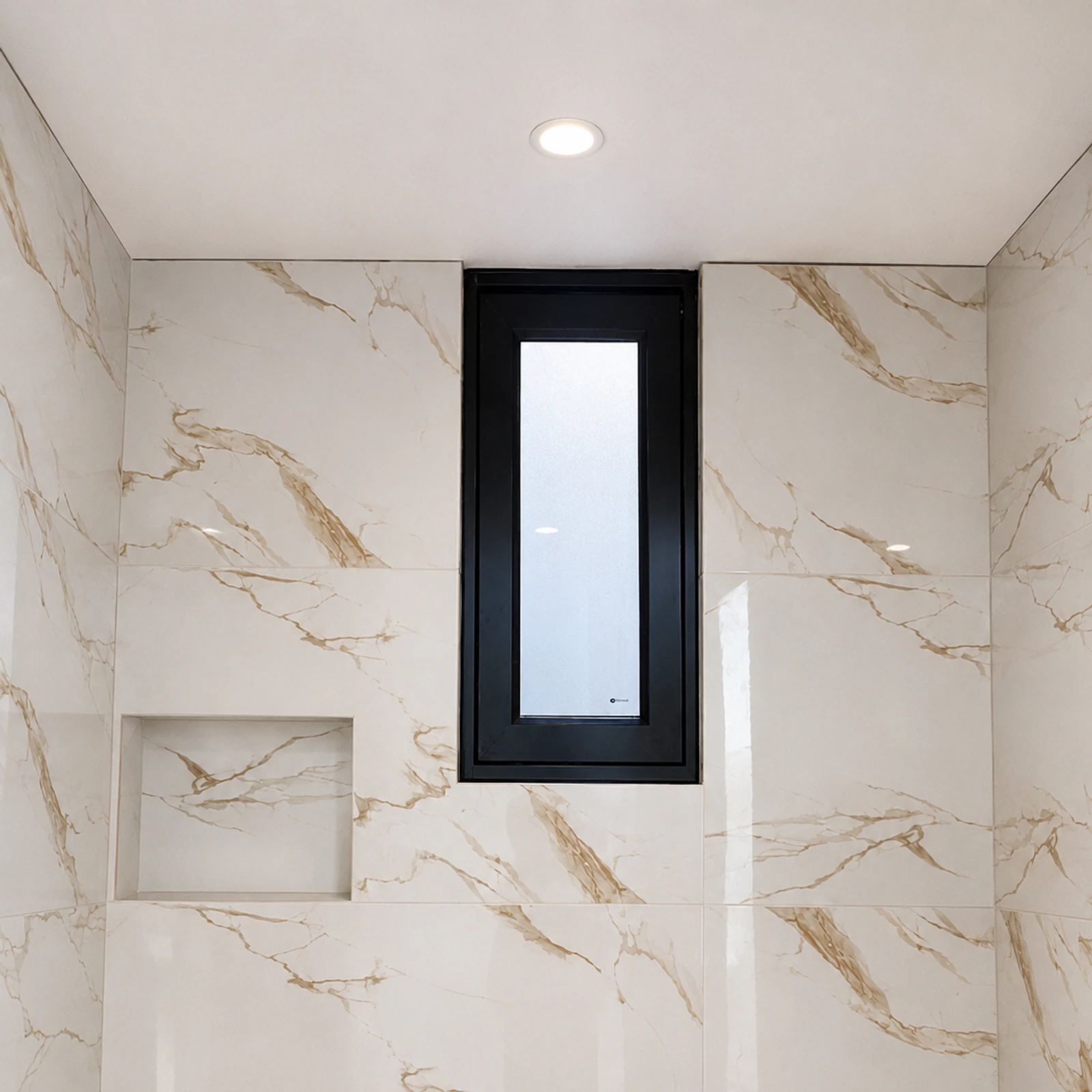 Ventana pivotante de aluminio negro terminada en un baño de revestimiento marmoleado