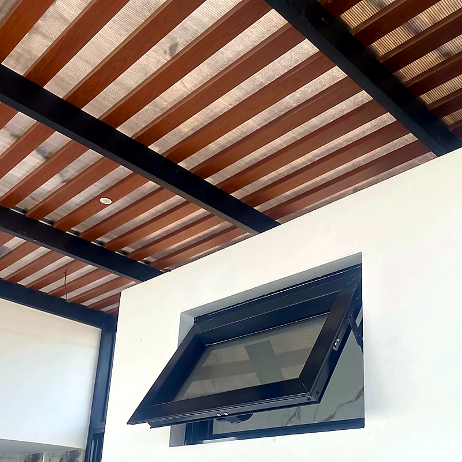 Ventana proyectante de aluminio negro abierta bajo un techo de vigas de madera